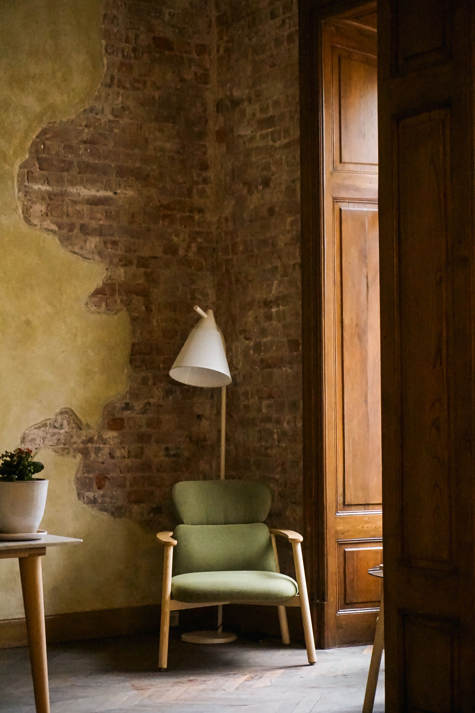 A stylish apartment interior featuring a green armchair, lamp, and rustic exposed brick walls.