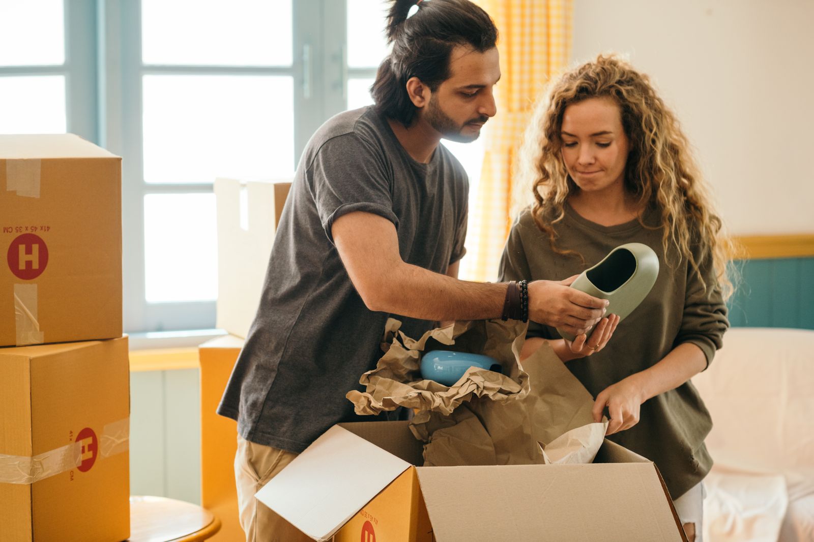 Crop ethnic man showing ceramic vase to girlfriend standing near pile of cardboard boxes with parchment after relocating to new house