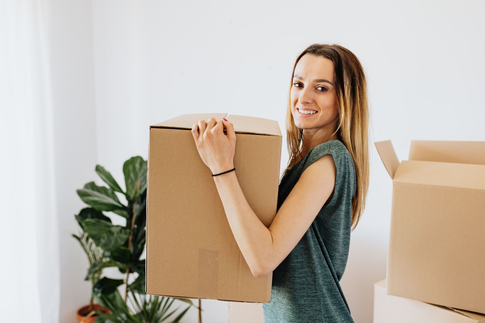 Side view smiling lady in casual green shirt carrying cardboard box while preparing for moving in new home and looking at camera happily