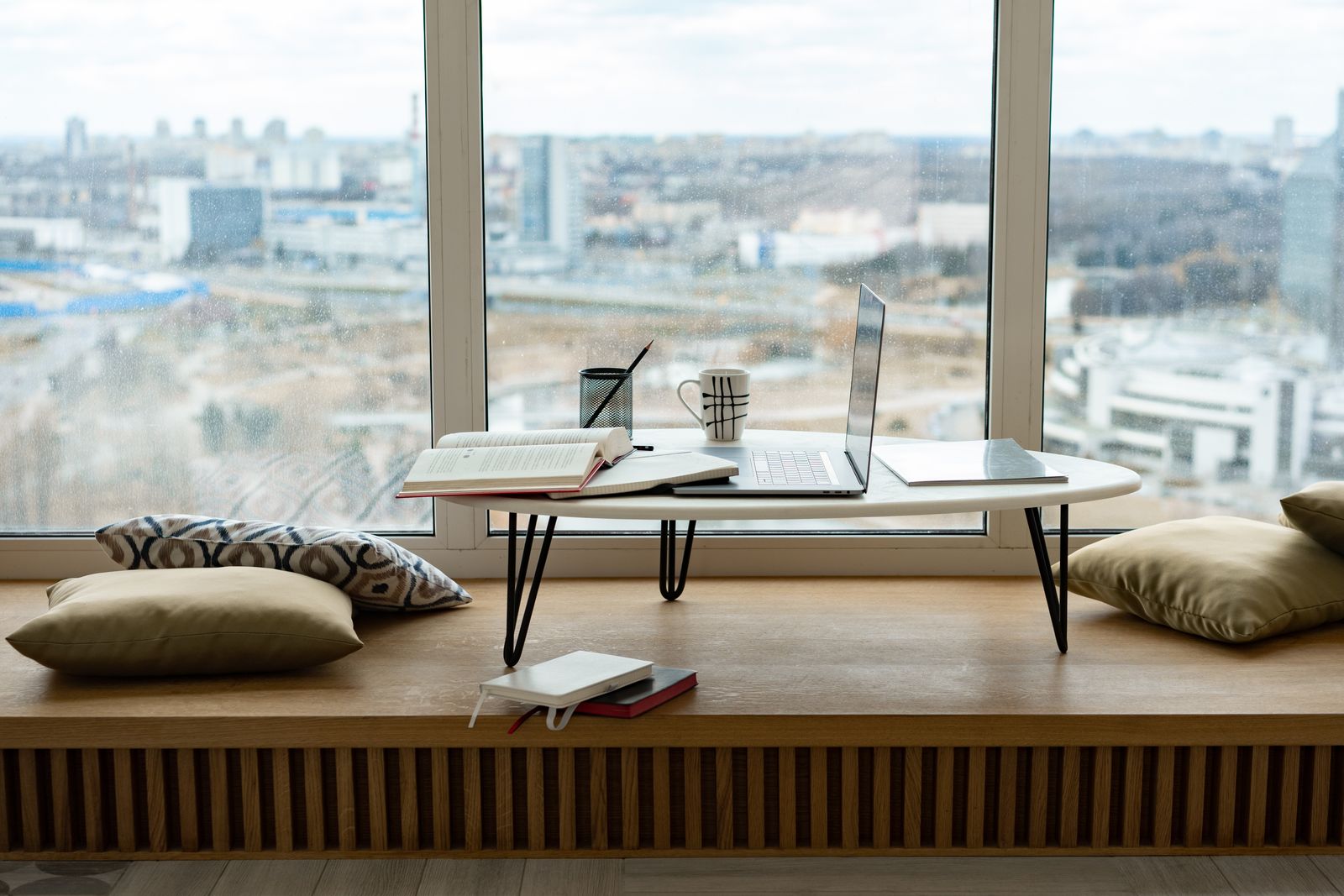 Minimalist workspace setup by a window with laptop, books, and cushions overlooking cityscape.